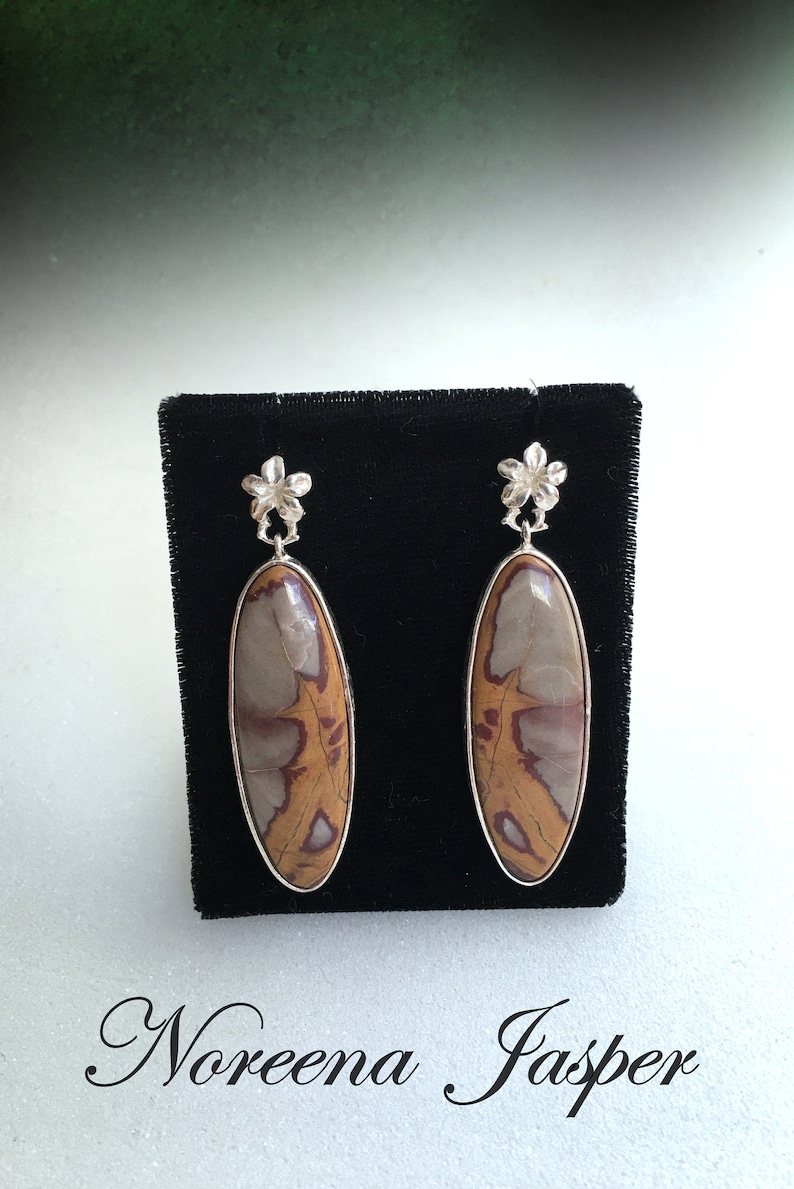 May include: A pair of silver earrings with a floral design and oval-shaped brown and white Noreena Jasper stones.