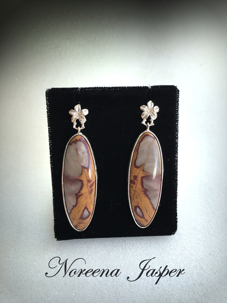 May include: A pair of silver earrings with a brown and white Noreena Jasper stone. The earrings have a flower design at the top.