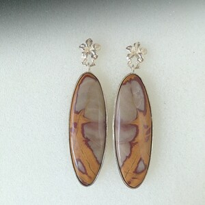May include: A pair of silver earrings with oval-shaped brown, white, and red jasper stones. The earrings have a flower-shaped design at the top.