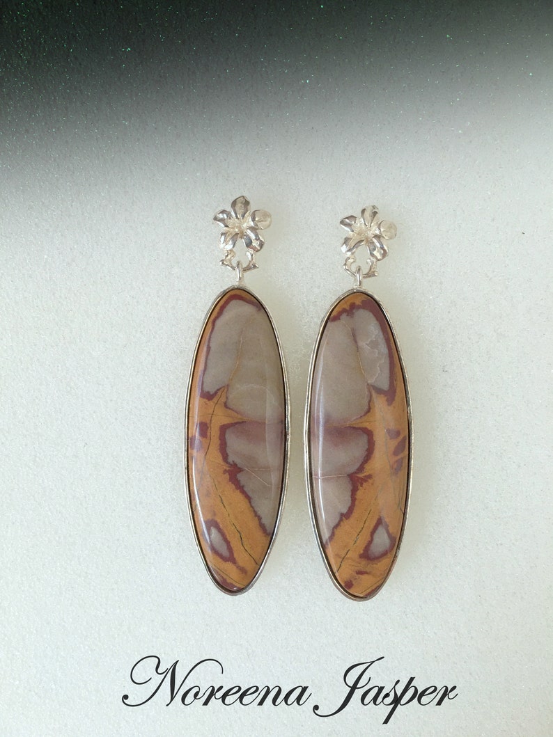 May include: A pair of silver earrings with oval-shaped brown, white, and red jasper stones. The earrings have a flower-shaped design at the top.