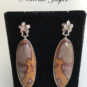 May include: A pair of silver earrings with a floral design and a brown, white, and red jasper stone. The earrings are displayed on a black velvet background.