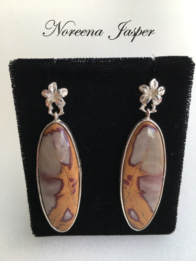 May include: A pair of silver earrings with a floral design and a brown, white, and red jasper stone. The earrings are displayed on a black velvet background.