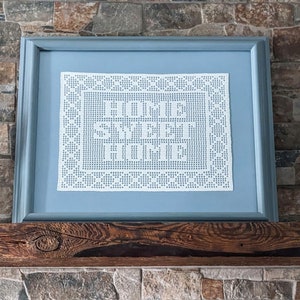 Könnte beinhalten: Ein gerahmtes Stück weißer Häkelarbeit mit den Worten "Home is where the heart is" in der Mitte. Der Rahmen ist hellblau und steht auf einem Holzmantel.