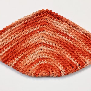 May include: A crocheted dishcloth in shades of orange and peach. The dishcloth is made with a simple stitch pattern and has a diamond shape.