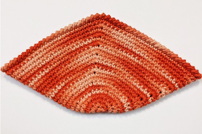 May include: A crocheted dishcloth in shades of orange and peach. The dishcloth is made with a simple stitch pattern and has a diamond shape.