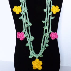 May include: A green crocheted necklace with pink and yellow flowers. The necklace is displayed on a black mannequin.