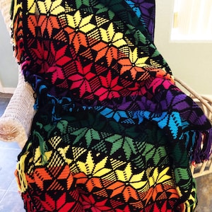 Peut inclure: Une couverture en crochet arc-en-ciel avec un fond noir et un motif en étoile. La couverture a des franges sur tous les côtés.