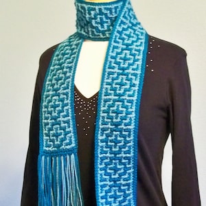 May include: A teal and blue crocheted scarf with fringe on a black mannequin. The scarf has a geometric pattern.