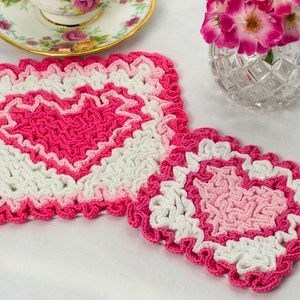 Può includere: Due tovagliette all'uncinetto a forma di cuore in rosa e bianco. La tovaglietta più grande è in primo piano e quella più piccola è dietro.