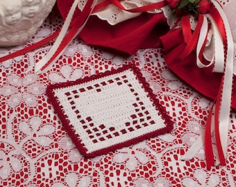 May include: A square, handmade crochet coaster with a white center and a red border. The coaster features an open heart design in the center and is placed on a white lace tablecloth. Red and white ribbons are in the background.