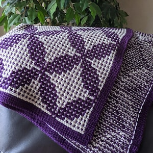 May include: A purple and white knitted throw blanket with a geometric pattern. The blanket is folded in half and is sitting on a purple couch.