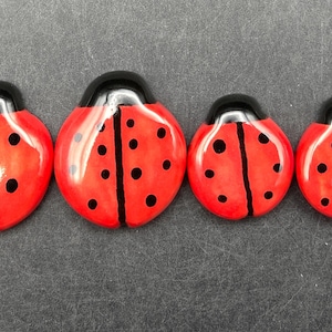 May include: Four red ladybug-shaped decorative items with black spots and black heads are arranged in a row. The ladybugs vary in size, with the largest on the left and the smallest on the right. The ladybugs are on a dark gray surface.