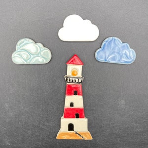 Puede incluir: Una figurita de faro de cerámica con rayas rojas y blancas, una banda negra y una luz amarilla. El faro está rodeado de tres figuritas de nubes de cerámica, una blanca, una azul y una azul claro.
