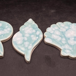 以下が含まれることがあります： さまざまなデザインの、貝殻の形をした3つのセラミックコースター。各コースターは、白の雲のような模様が入った、明るいティール色です。コースターは暗い表面に配置されており、そのユニークな形と質感を際立たせています。
