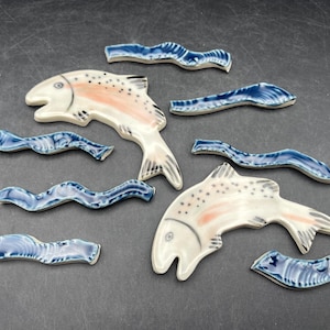 May include: Ceramic fish and wave-shaped tiles. The fish are white with pink and black accents. The wave tiles are blue and white. The items are arranged on a dark grey surface.
