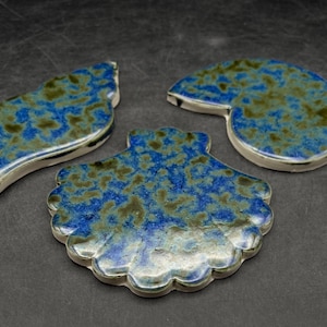 以下が含まれることがあります： 葉、貝殻、オウムガイの形をした3つのセラミック装飾品。青と緑のまだら模様の釉薬と白い縁が特徴です。オブジェクトは濃い灰色の表面にあります。