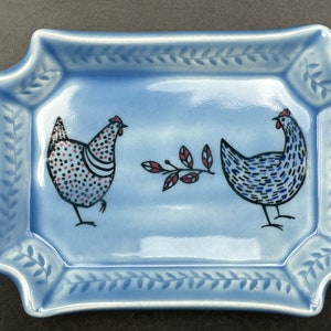 May include: A light blue ceramic dish with a decorative border of leaves. Two stylised chickens are painted on the dish, one in black, white, and red, and the other in blue and white. A small branch with red berries is painted between the chickens.