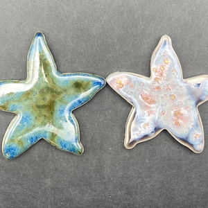 May include: Two ceramic starfish decorations. One is blue, green, and gold, the other is white, pink, and blue. Both have a glossy finish and a silver-colored edge. The starfish are on a gray background.