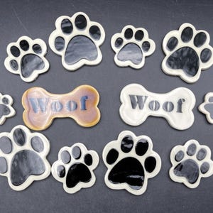Puede incluir: Una colección de decoraciones de cerámica con temática de perros, que incluyen huellas de patas y piezas en forma de hueso. Las huellas de patas son blancas con almohadillas negras, y las piezas en forma de hueso tienen la palabra "Woof".