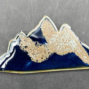 May include: A ceramic mountain-shaped decorative piece. The piece features a dark blue base with a textured, light brown and white center, and a white and gold trim. The mountain range design is a unique home decor item.