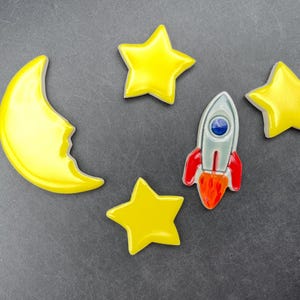 May include: A selection of space-themed decorations, featuring a yellow crescent moon, three yellow stars, and a silver, red, and orange rocket. The items are displayed on a dark grey surface.