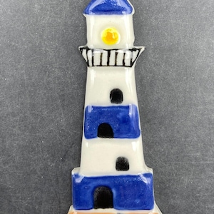 Puede incluir: Figura de faro de cerámica con techo azul, cuerpo blanco y rayas azules. El faro tiene una luz amarilla y ventanas negras. La base es naranja. Un objeto náutico decorativo.
