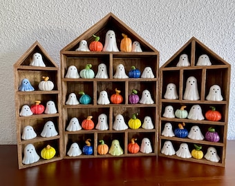 Puede incluir: Tres estantes de madera con forma de casa que contienen una colección de pequeñas figuras de cerámica. La muestra presenta fantasmas blancos con ojos y bocas negras, junto a calabazas en miniatura de colores naranja, amarillo, azul, verde y morado.