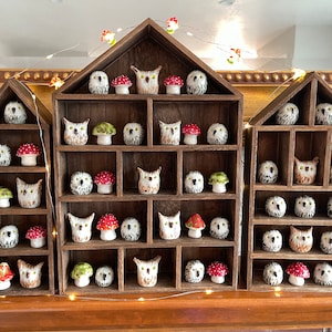 May include: A wooden shelf unit with a house-shaped design, featuring multiple compartments filled with small ceramic owl figurines. Each owl is decorated with white and black markings, and some have red and green mushroom accents.