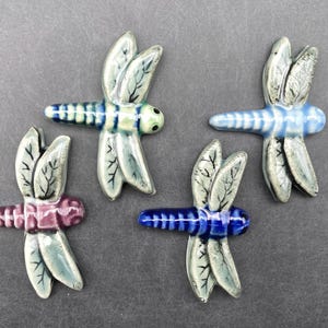 May include: Four ceramic dragonfly ornaments in blue, green, and pink. Each dragonfly has detailed wings and a striped body. The wings are a light green colour with dark veins. The bodies are striped in various colours.