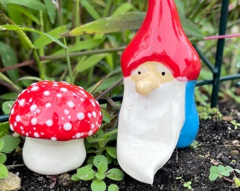 Może przedstawiać: Ceramiczna figurka gnoma z czerwonym kapeluszem, białą brodą i niebieskimi ubraniami stoi obok ceramicznej figurki grzyba w kolorze czerwonym i białym.