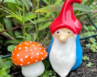 Może przedstawiać: Ceramiczna figurka gnoma z czerwonym kapeluszem, białą brodą i niebieską koszulą stoi obok ceramicznego grzyba w pomarańczowe i białe kropki.