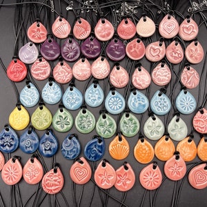 Puede incluir: Una colección de 50 colgantes de cerámica con varios diseños y colores, colgados de cordones negros. Los colgantes tienen forma de lágrima y presentan patrones florales, geométricos y abstractos.