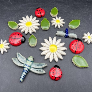 May include: A collection of ceramic garden ornaments including white daisies with yellow centers, green leaves, and red ladybugs with black spots. A blue and white dragonfly is also included.