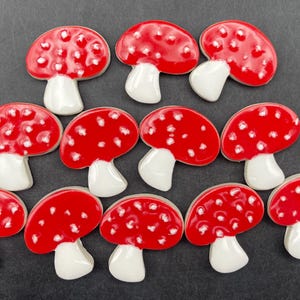 May include: A collection of ten red and white mushroom-shaped ceramic decorations. Each mushroom has a red cap with white spots and a white stem. The mushrooms are arranged on a dark gray surface.