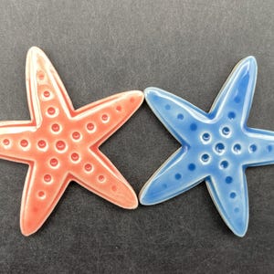 May include: Two ceramic starfish, one coral and one blue, each with five points and a textured surface. The starfish are arranged side by side against a dark background, evoking a coastal or nautical theme.