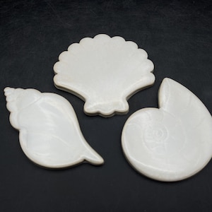 以下が含まれることがあります： 3つの白い貝殻型の装飾品。ホタテ貝、巻貝、オウムガイの貝殻が含まれています。貝殻は暗い表面に配置されています。貝殻は滑らかで光沢のある素材で作られています。