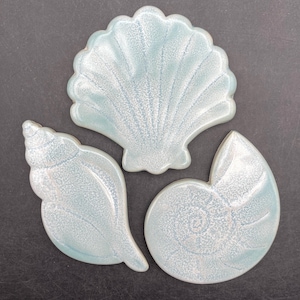 Op de afbeelding: Drie keramische schelpen decoraties in blauw- en wittinten. De set bevat een sint-jakobsschelp, een kauri schelp en een nautilusschelp. De schelpen hebben een glanzende afwerking en zijn gerangschikt op een donkergrijze ondergrond.