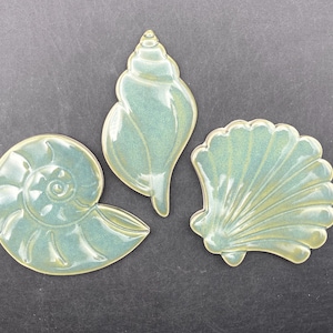 以下が含まれることがあります： 海の色と黄色の色合いの3つのセラミック貝殻装飾。 セットには、オウムガイ、コンク、ホタテ貝が含まれており、すべて光沢のある仕上がりです。 貝殻は濃い灰色の表面に配置されています。
