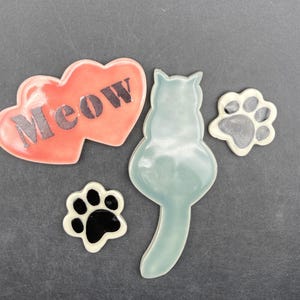 May include: A selection of ceramic cat-themed decorations. Features a heart with the word "Meow", a cat silhouette, and two paw print designs. The pieces are in shades of red, blue-green, black, and white.