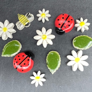 Könnte beinhalten: Eine Sammlung von Keramik-Gartendekorationen, darunter weiße Gänseblümchen mit gelben Zentren, grüne Blätter, rote Marienkäfer mit schwarzen Punkten und eine gelb-schwarze Biene. Ideal für die Gartendekoration.
