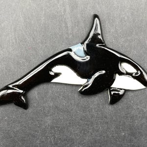 May include: A glossy, black and white orca figurine with a light blue accent on its back. The orca is depicted in a swimming pose, with its dorsal fin raised. The figurine is likely made of ceramic or a similar material.