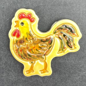 May include: A ceramic rooster figurine with a yellow base and a detailed design. The rooster has a red comb and wattle, with a yellow body and a colorful tail. The figurine is likely a decorative item.