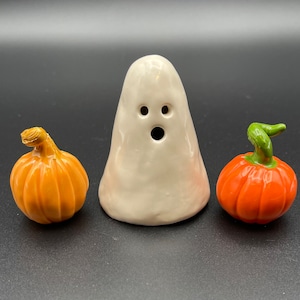 May include: A white ceramic ghost figurine with two small circular eyes and a larger circular mouth, flanked by two small pumpkins. The pumpkin on the left is light orange with a tan stem, and the pumpkin on the right is bright orange with a green stem.