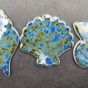 以下が含まれることがあります： 3つの貝殻型のセラミック装飾品。青と緑のまだら模様に釉薬が施されています。形状には、ホタテ貝、オウムガイ、巻貝が含まれます。