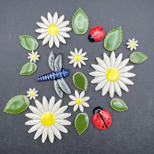 Op de afbeelding: Een verzameling keramische tuindecoraties, waaronder witte madeliefjes met gele centra, groene bladeren, rode lieveheersbeestjes en een blauwe libelle. De stukken zijn gerangschikt op een donkergrijze ondergrond.