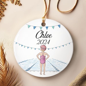 Puede incluir: Adorno redondo de cerámica blanca con una ilustración de nadadora. El adorno presenta el nombre "Chloe" y el año "2024" en escritura negra. Una nadadora con traje de baño y gorro rosas está frente a una piscina azul.