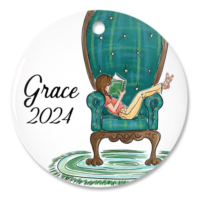 May include: A round, white ornament with the text "Grace 2024". The ornament features an illustration of a person reading a book while relaxing in a teal armchair with a green plaid pattern. The person is wearing yellow pants and a pink shirt.