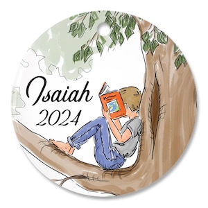 May include: A round ceramic ornament featuring a watercolor illustration of a person sitting in a tree reading an orange book. The text Isaiah and 2024 are printed in black script and serif font on the left side of the white background.