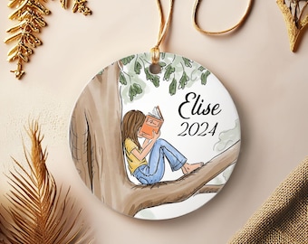 Puede incluir: Adorno navideño redondo de cerámica que representa a una niña leyendo un libro en la rama de un árbol. La niña lleva una camiseta amarilla y vaqueros azules. El adorno tiene el nombre "Elise" y el año "2024".