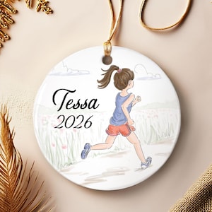 May include: A round white ceramic ornament featuring a printed illustration of a person with a ponytail running through a field. The text "Tessa" and "2026" is printed in black script and serif font. A gold string is attached to the top hole.